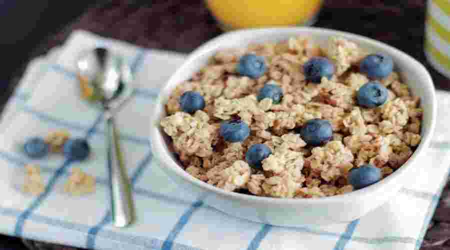 A close-up shot of a bowl of cereal with blueberries A close-up, high-angle shot of a white bowl filled with granola or muesli, generously topped with fresh blueberries. A silver spoon rests on a blue and white checkered napkin next to the bowl, with a few extra blueberries scattered nearby.