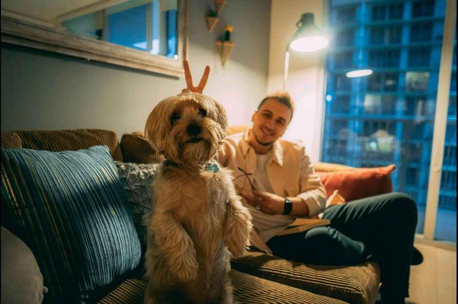 Man and his dog on a couch A man sits on a brown corduroy couch, smiling at the camera. In front of him, a small, shaggy brown dog is standing on its hind legs, with a small toy or treat balanced on its head, looking directly at the camera. The scene is illuminated by a warm lamp light, creating a cozy atmosphere.