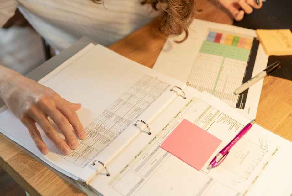 Planning and Organizing Documents An overhead shot of a person's hands reviewing papers and a ruler in an open three-ring binder, with a pink sticky note on one page and various documents and a pen scattered on a wooden table