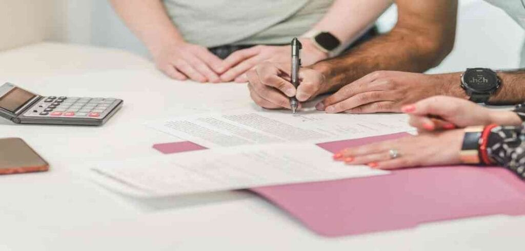 Signing a Contract Three people's hands are visible as one person signs a document on a white table with a pen, near a calculator, a phone, and pink paper.